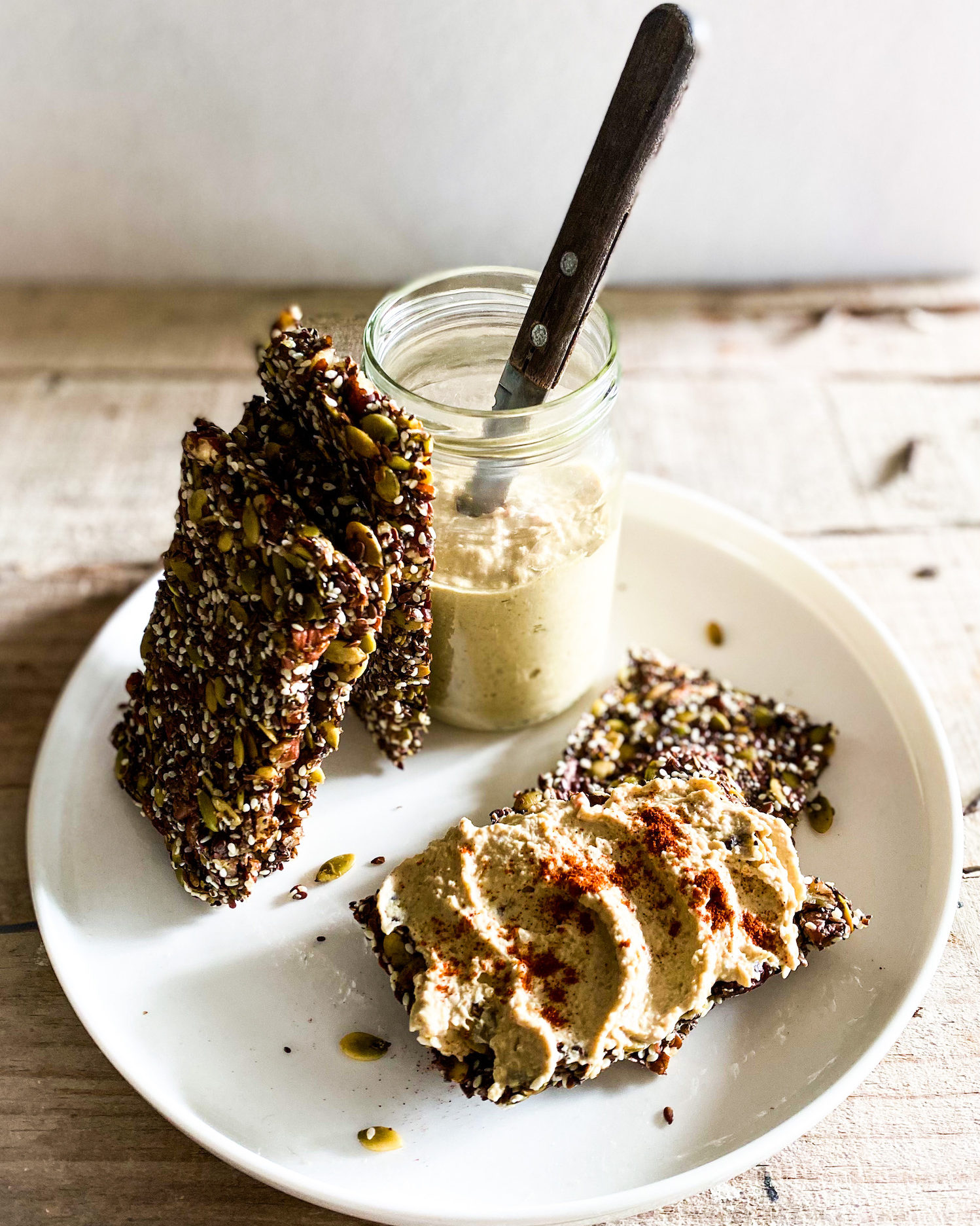 nuts and seeds crackers stacked against a jar of hummus and 1 cracker with hummus spread on it